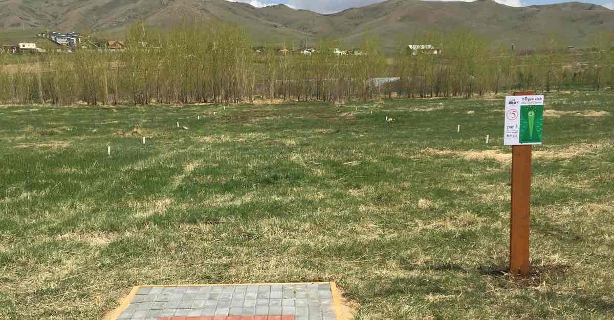 The end of a disc golf tee pad and a tee sign in an open field with mountains beyond