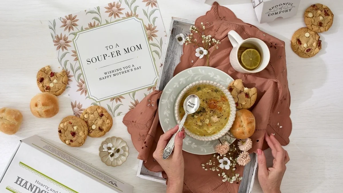 An overhead view of a Mother’s Day table setting with a bowl of soup being held and stirred, surrounded by cookies, bread rolls, a teacup with lemon, floral accents, and a card that reads “To a Souper Mom — Wishing You a Happy Mother’s Day.”