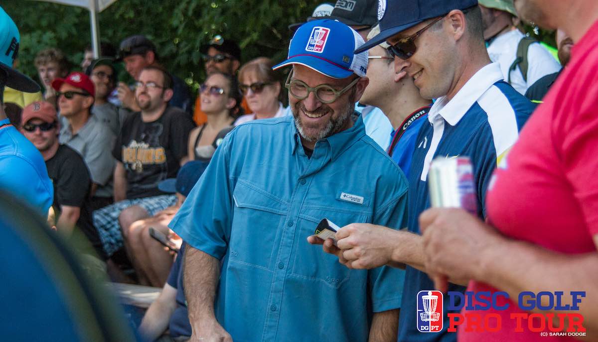 A man in circle rim glasses laughing in the middle of a crowd at a disc golf event