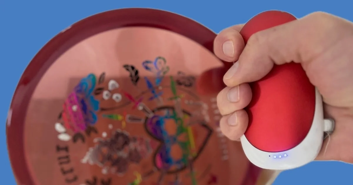 A hand holding a battery-powered handwarmer in front of a disc golf disc on a blue background