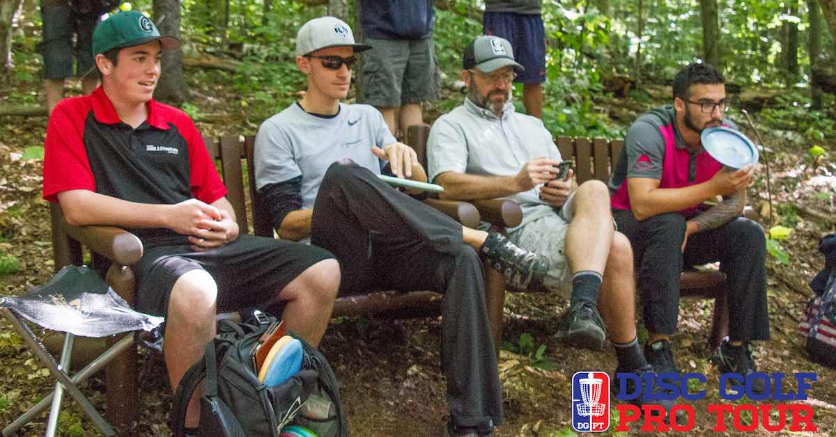 Four men sit on a bench in the woods with discs and disc golf attire