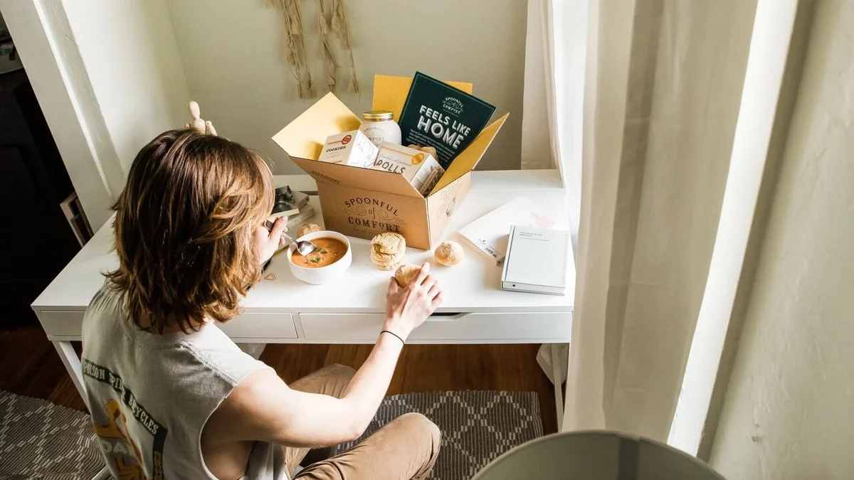 Person sitting at a desk with an open Spoonful of Comfort gift box, enjoying a bowl of soup while reading a book, with tea and a handwritten note nearby.