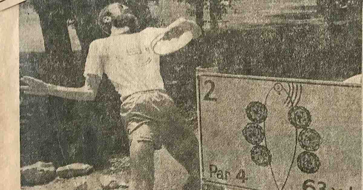 Black and white photo of an older man, "Steady" Ed Headrick, throwing a Frisbee into an original disc golf basket as a young man observes