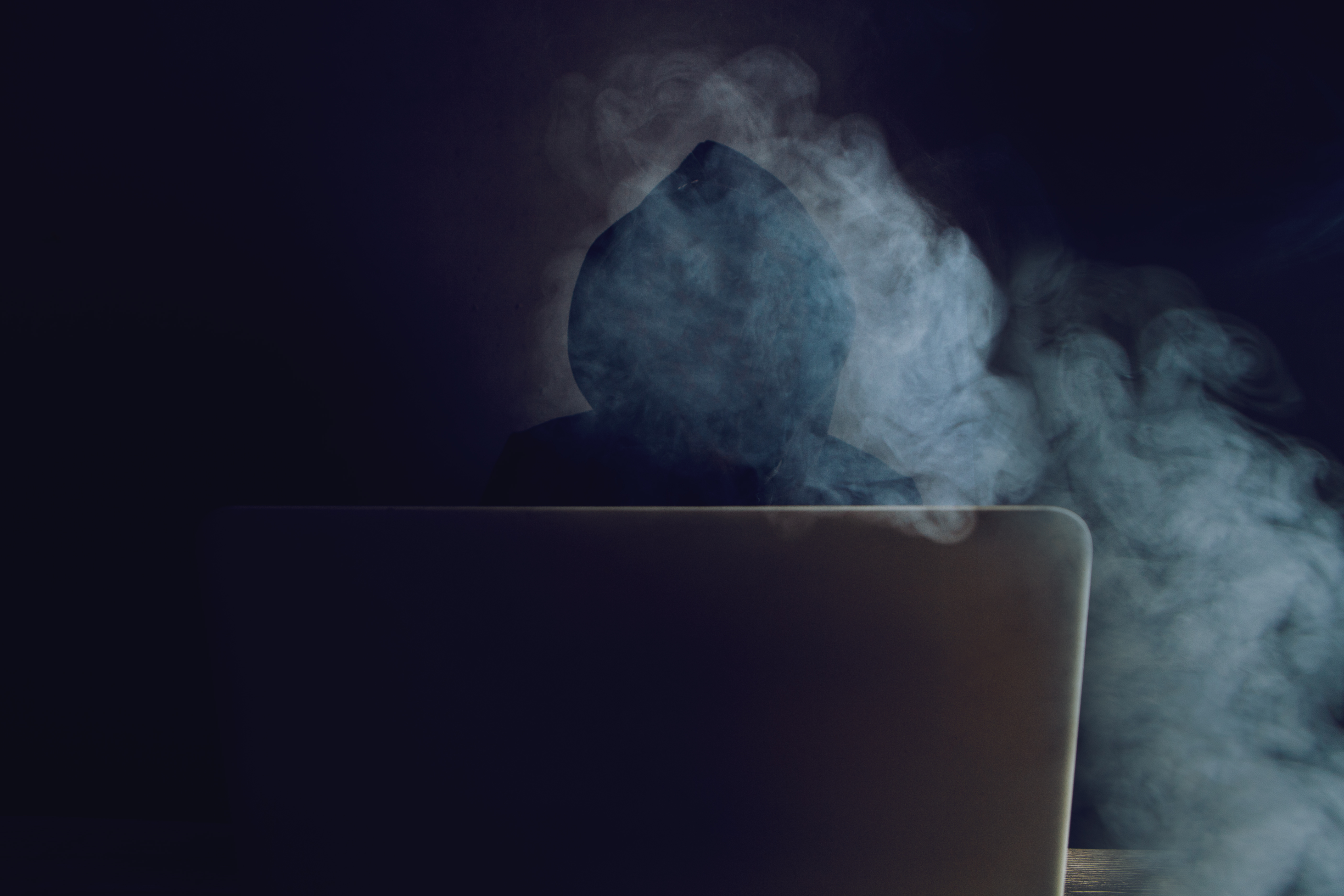 Person in hoodie sitting in front of laptop with smoke