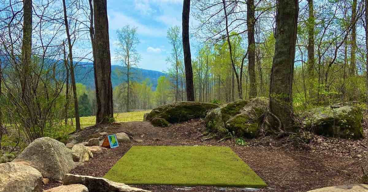 A turf disc golf tee pad in a somewhat wooded area. Open field and rolling mountains beyond.