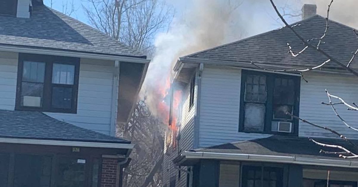 Upper stories of two house built close to one another. The right house has fire and smoke coming out of it.