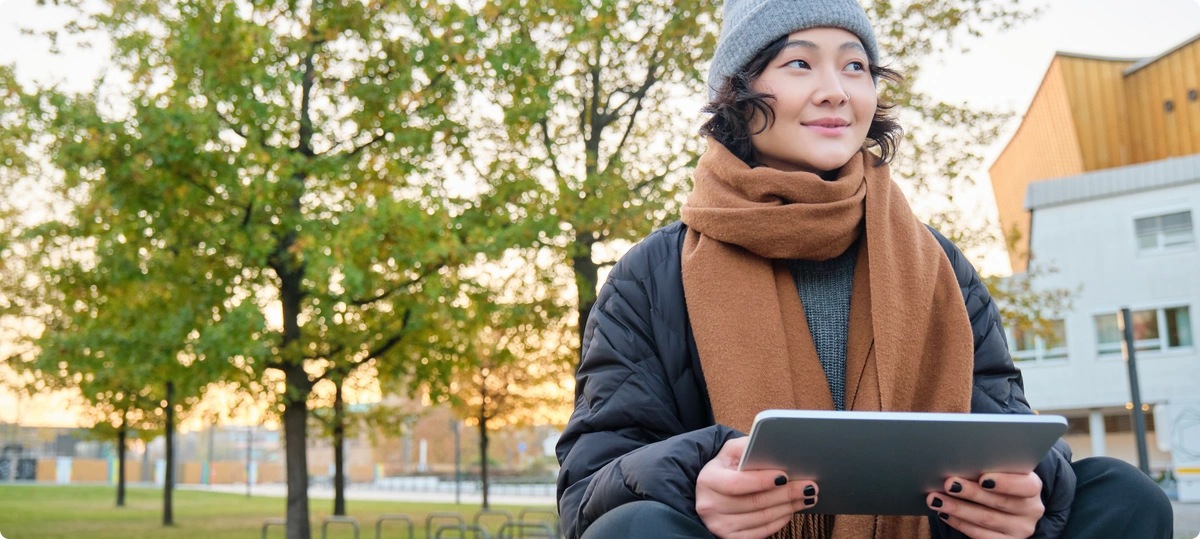 A woman dressed in warm clothing holding a tablet while standing outside with a tree and building behind her