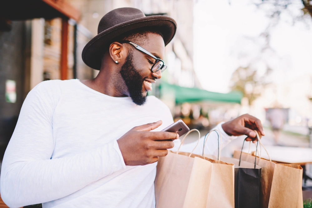 Man wearing a brown hat, white t-shirt and glasses holds his phone while sitting looking in brown paper shopping bags