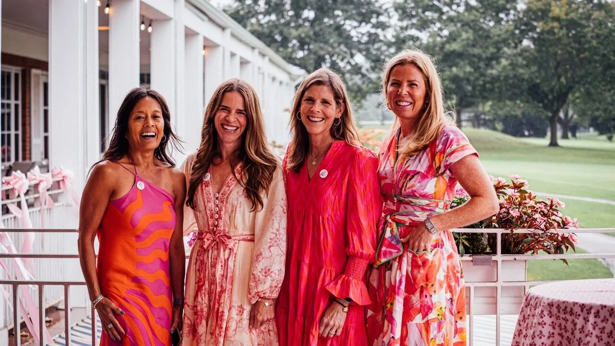 Four women wearing vibrant pink and floral dresses stand together smiling at an outdoor event decorated with pink accents.