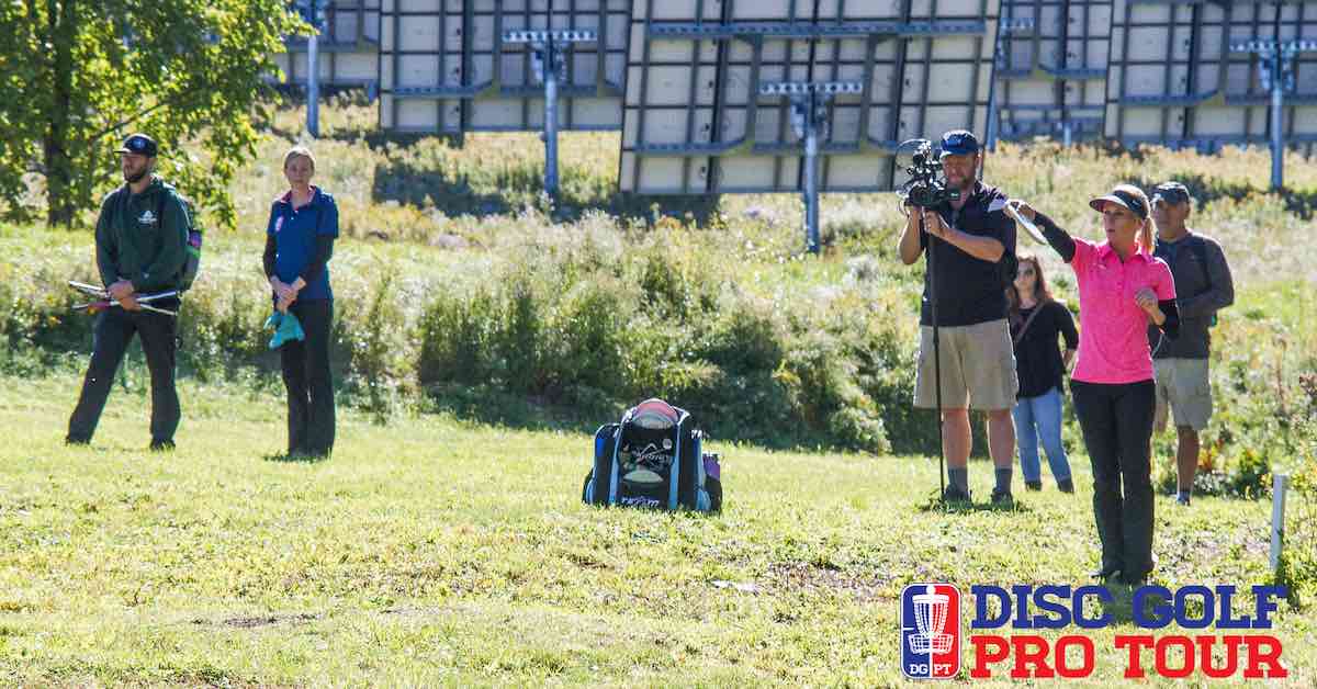 A woman in a pink shirt lines up a disc golf throw in an open green area. A cameraman catches the scene on film