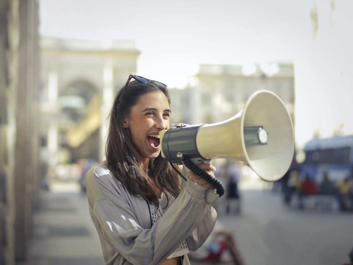 A female brand advocate is on a sidewalk holding a bullhorn. The woman is excited and smiling as she yells out her messages.