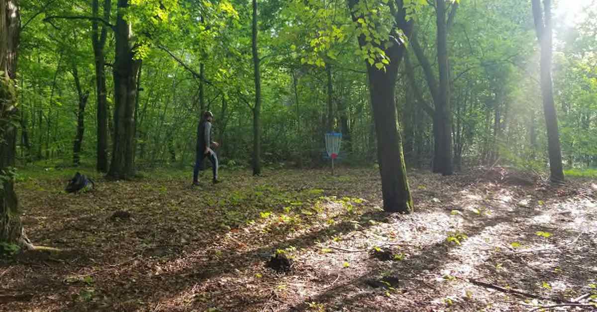 A man walks to a blue disc golf basket in the woods