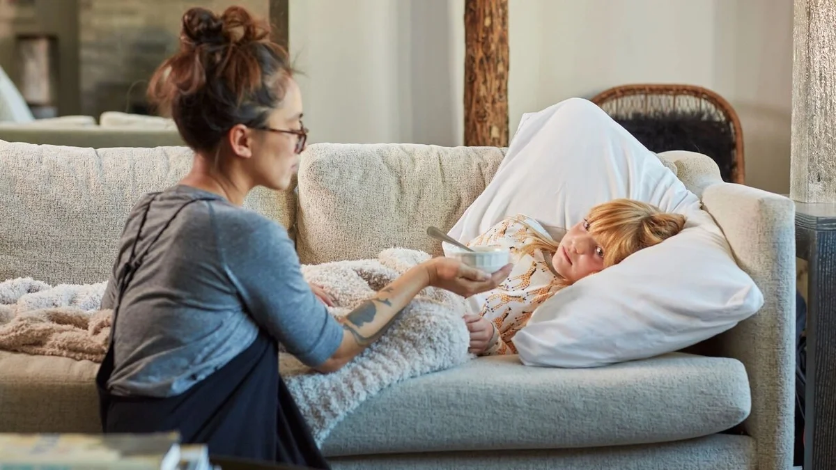 Adult sitting beside a child resting on a couch under a cozy blanket, offering a bowl of chicken soup while the child lies on a pillow, creating a caring, comforting scene.