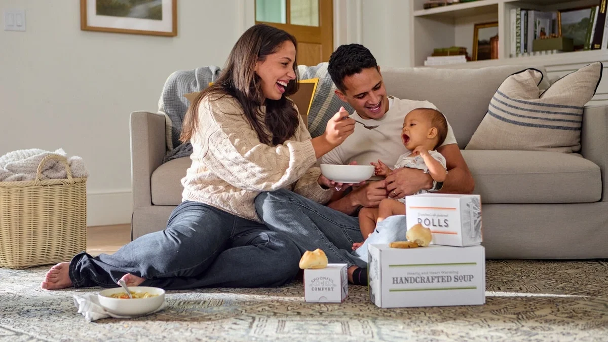 Parents sitting on the floor with their smiling baby, sharing a bowl of soup and a joyful moment together, with Spoonful of Comfort soup and rolls boxes nearby.