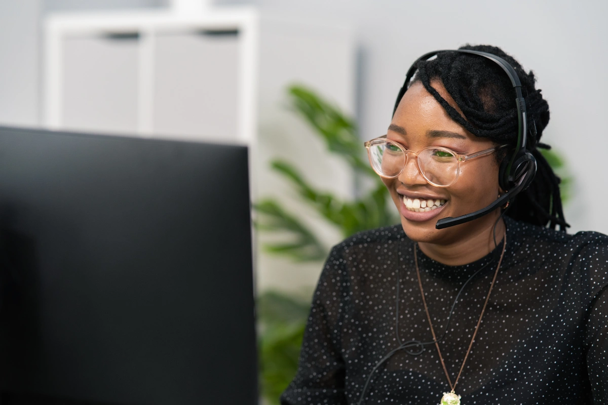 Black professional woman at a service desk, wearing a headset, working on a computer with a blurred office background.