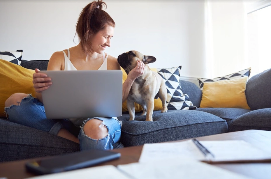 A woman sits cross-legged on a couch with a computer in her lap holding her French Bulldog's chin