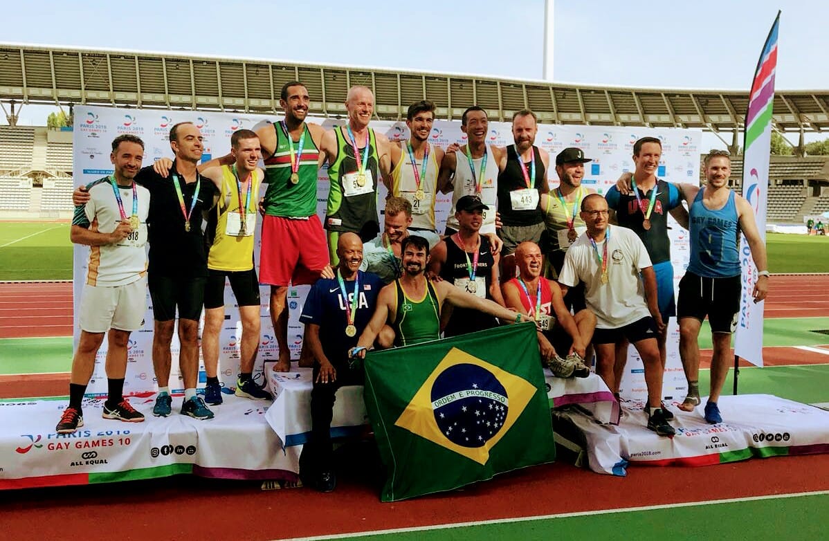 Group of men with medals around their necks on a podium smiling. One man is holding the Brazil flag in front of the group.