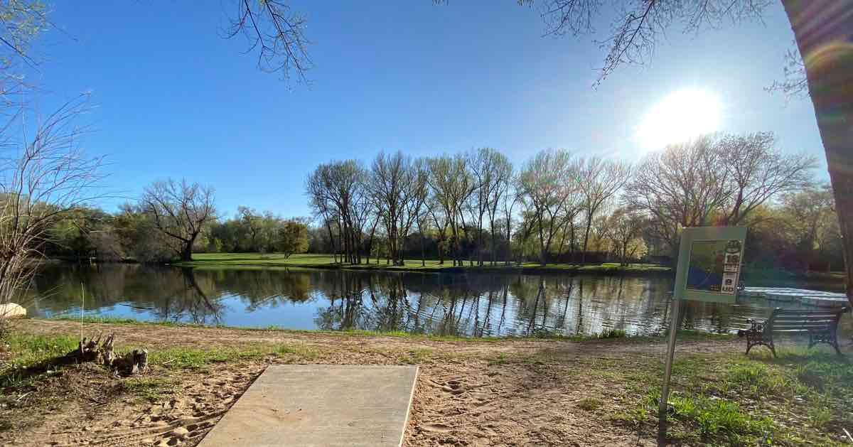 A concrete disc golf tee pad leads immediately to a pond with a line of trees guarding an open area of grass beyond