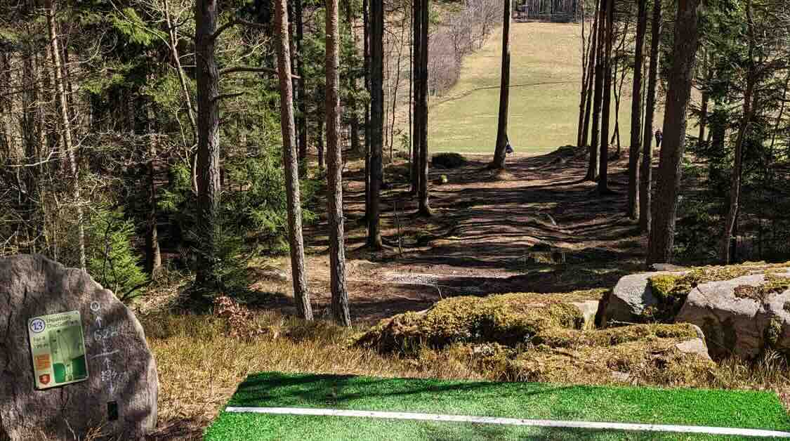 The end of a turf disc golf tee pad leads to a fairway that slopes through woods before turning into an open field