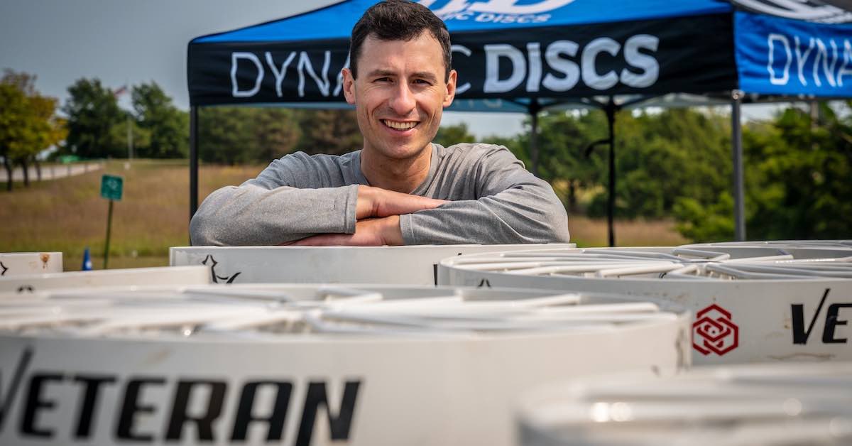 A man leans on the top of a disc golf basket with rows of other baskets in front of him