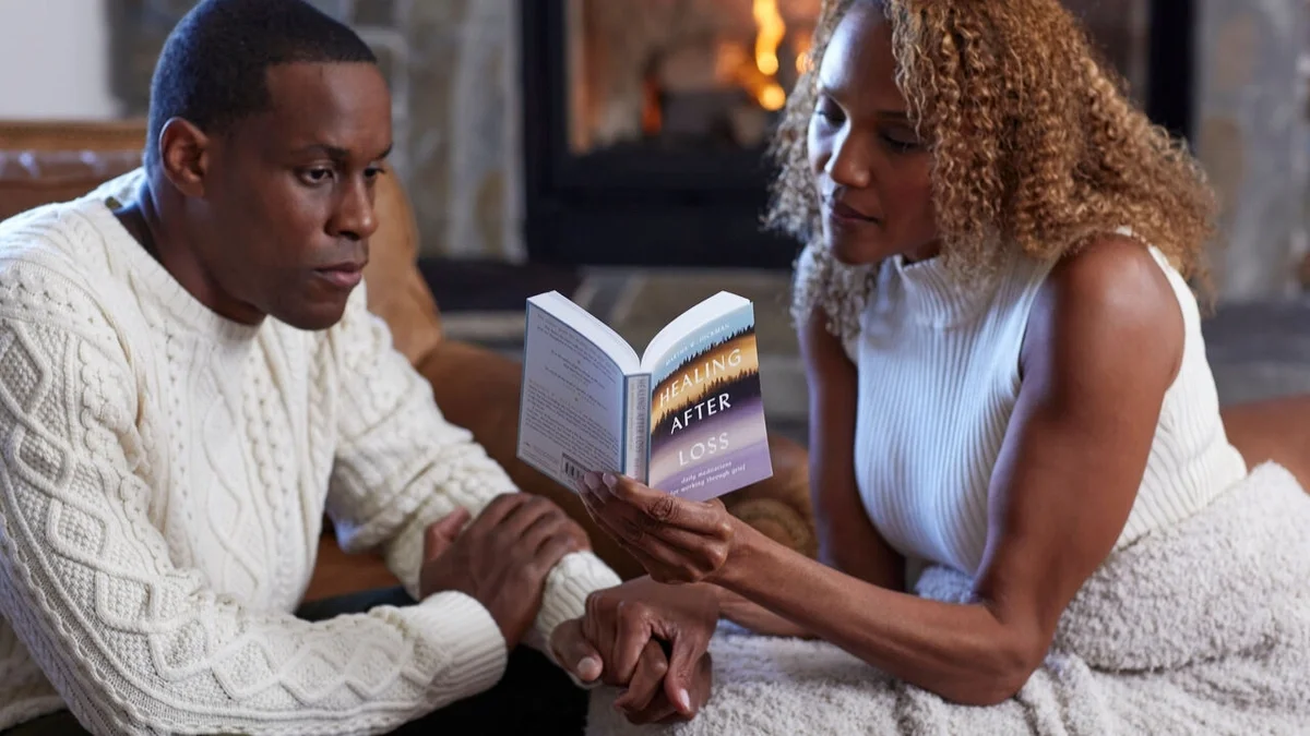 Two people sitting together in front of a fireplace, holding hands while one reads from the book Healing After Loss, sharing a quiet moment of comfort and support.