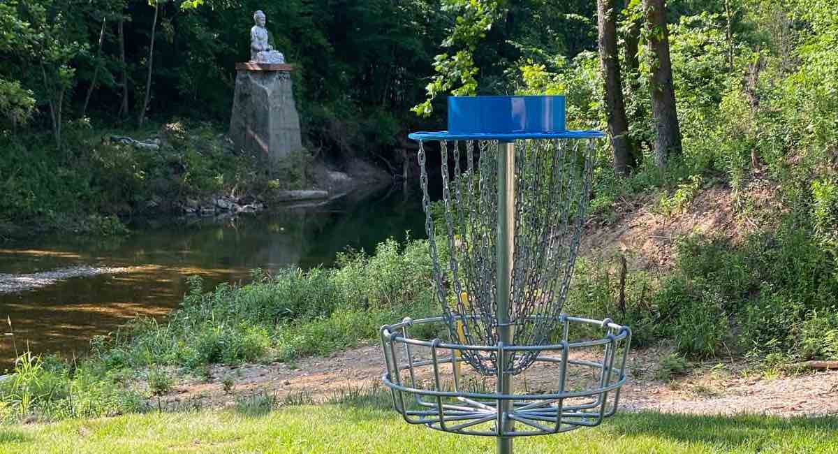 A blue disc golf basket in the foreground and a large Buddhist statue over water in the background