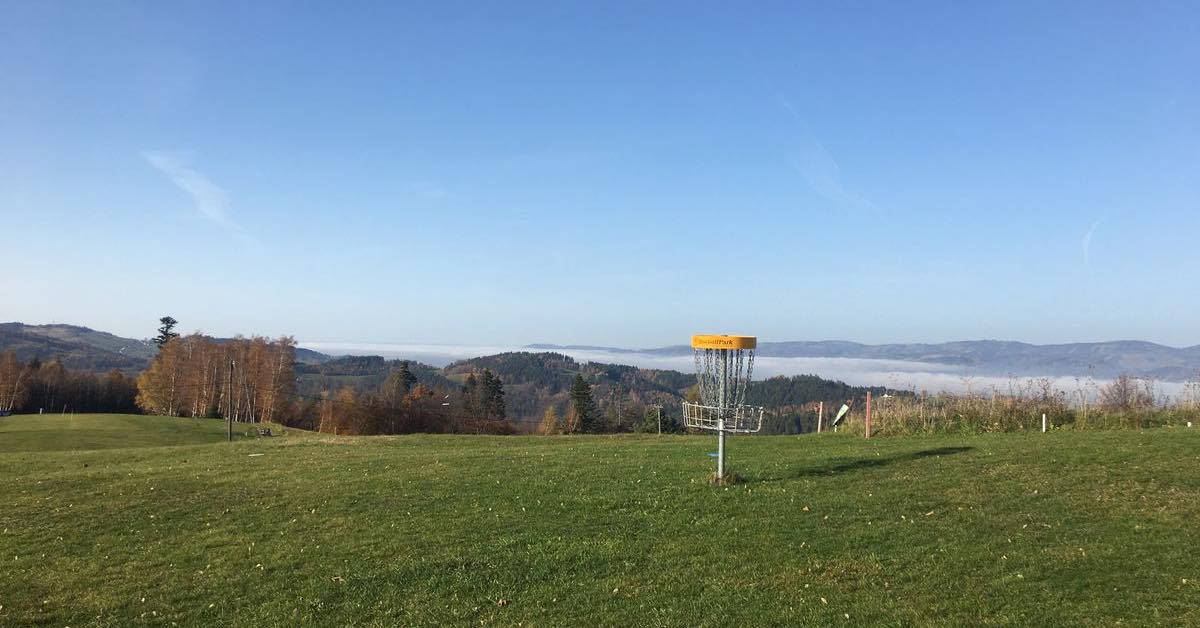 A disc golf basket on a hill covered in short green grass with mountains covered in cloud beyond