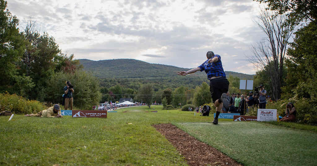 View from behind as a disc golfer in ablue shirt releases a disc down a very downhill fairway in the mountains