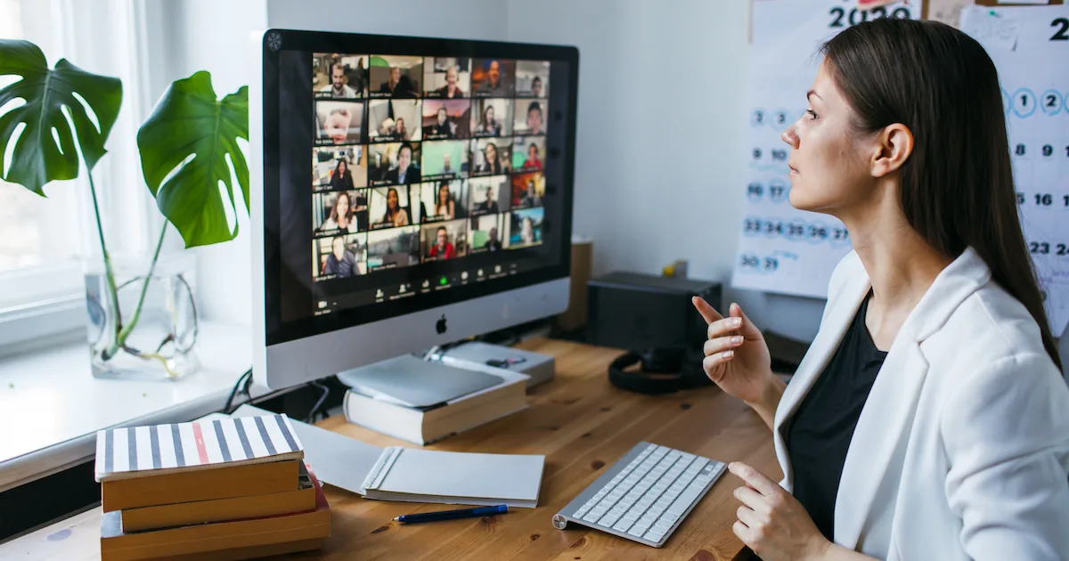 A young woman with brown hair and a white blazer participating in a virtual meeting with numerous participants.