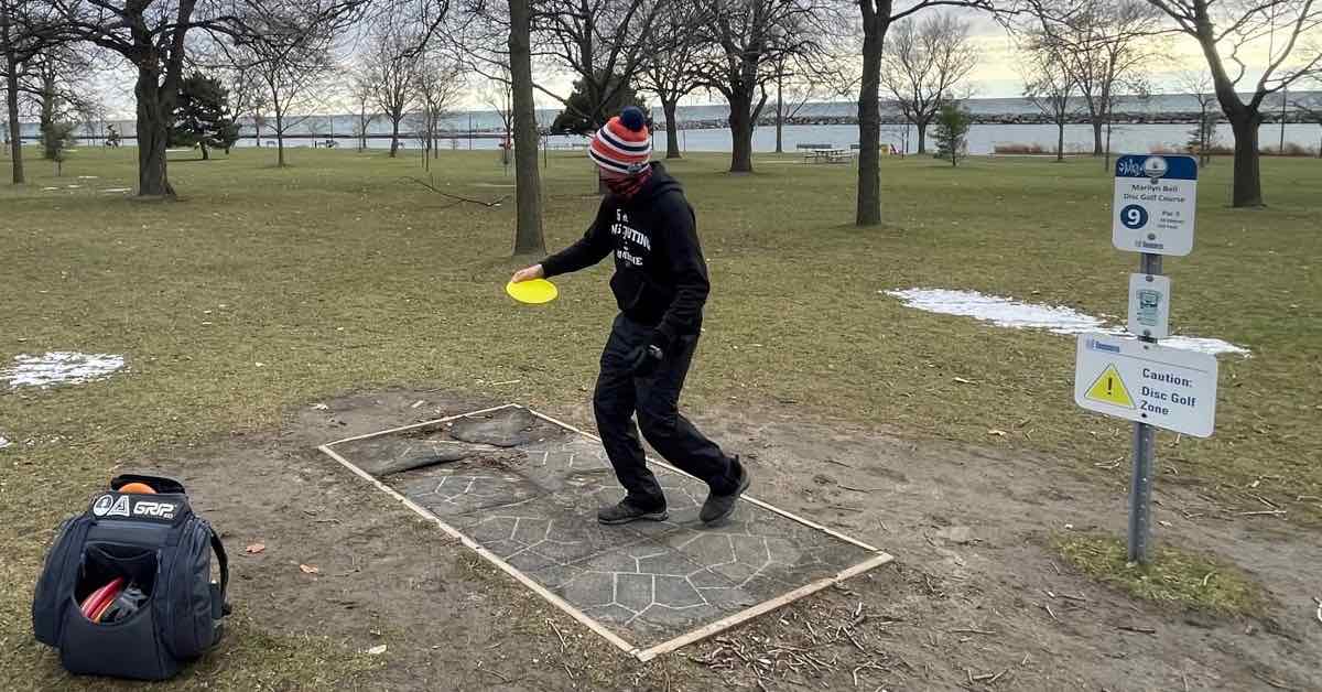 A man in winter clothes running up to throw a disc from a tee with park and waterfront in background