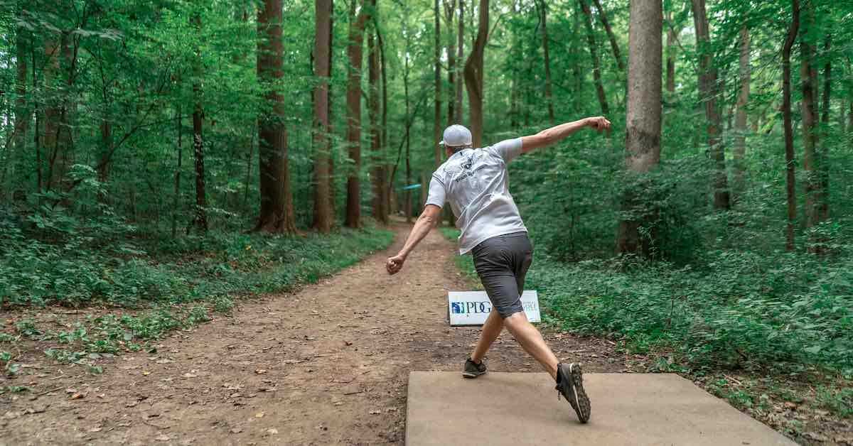 A disc golfer releasing a disc down a tightly wooded fairway