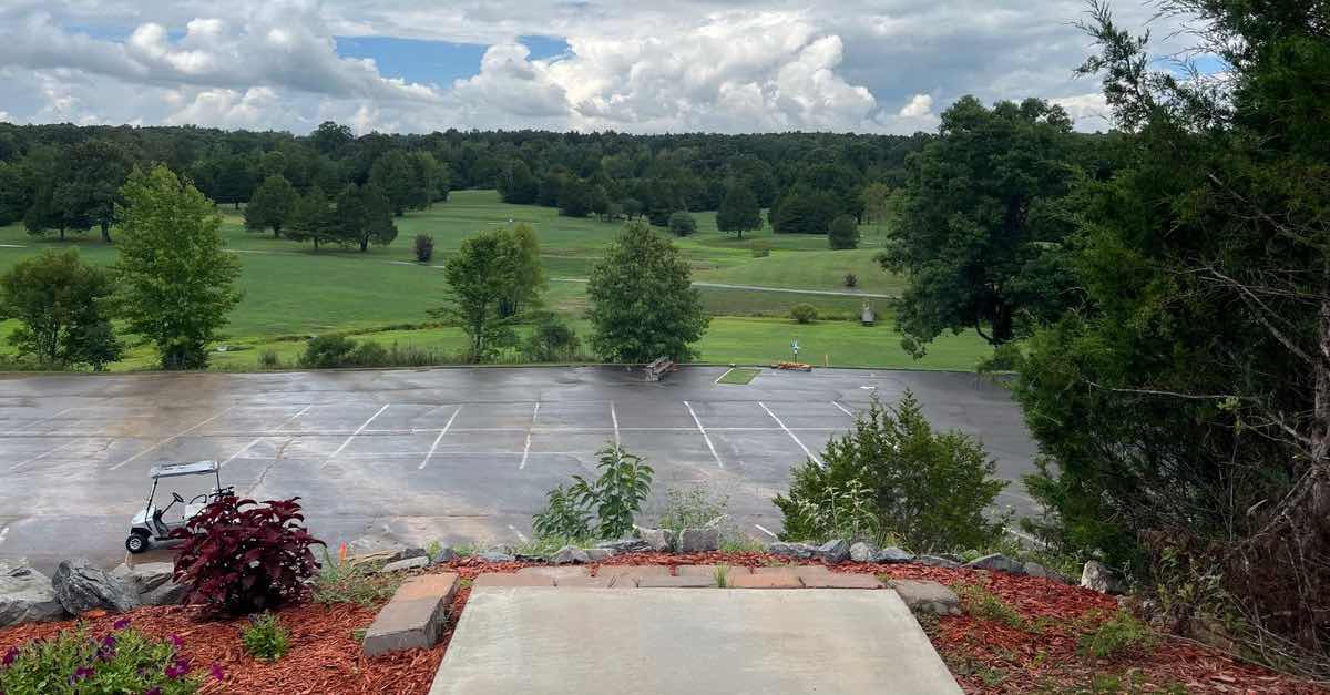 A concrete disc golf tee pad leads to a view over a parking lot and a manicured golf course space beyond