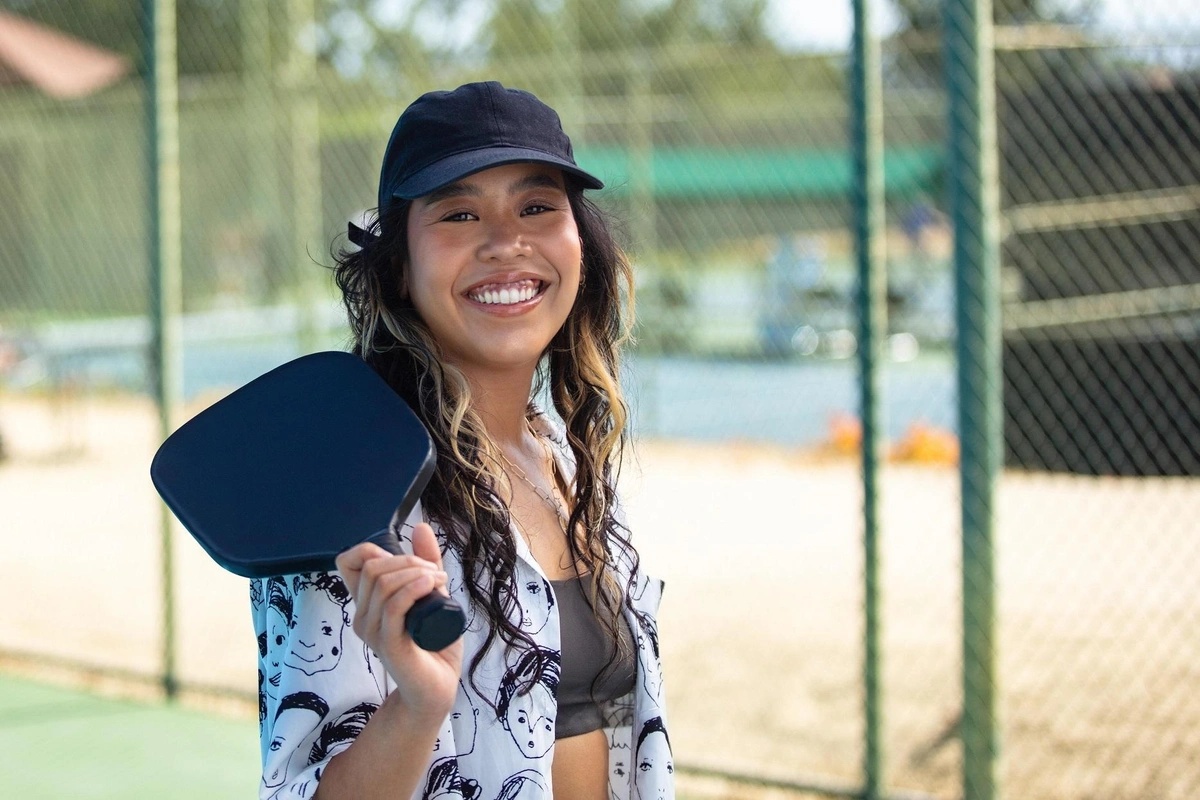 Woman smiling while holding a paddle and wearing a pickleball hat