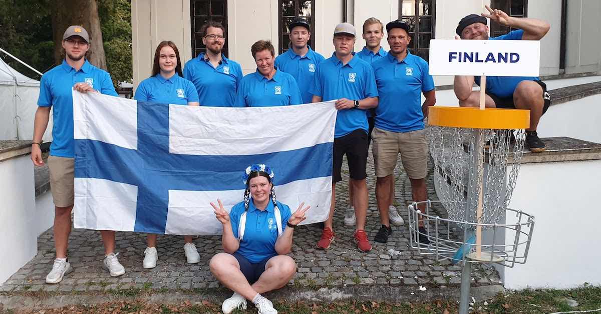 A group in Finland flag colors hold up a Finnish flag near a disc golf basket with a sign that says "Finland"