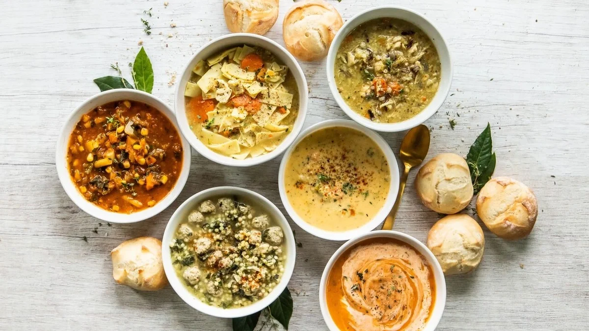 Overhead view of six bowls of assorted soups arranged with fresh rolls and green herbs on a light wooden surface.