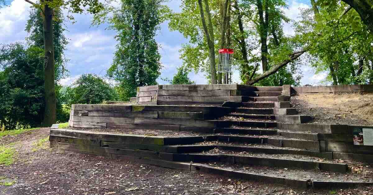 A red-banded disc golf basket on a tiered wooden structure with steps