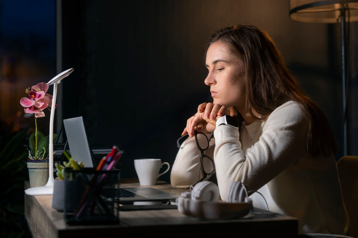 A woman sitting at a desk late at night, appearing thoughtful and focused while working on a laptop, with a cup of coffee and office supplies nearby.