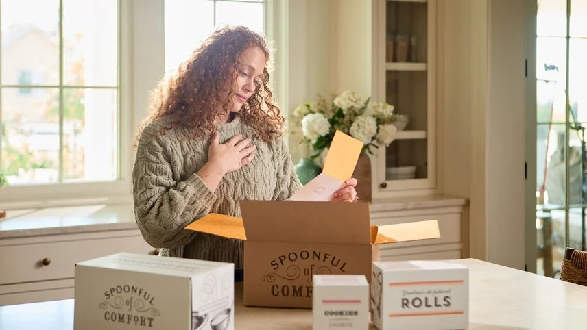Woman smiling as she reads a yellow greeting card after opening a Spoonful of Comfort gift box, with packaged soup, rolls, and cookies on the kitchen counter.
