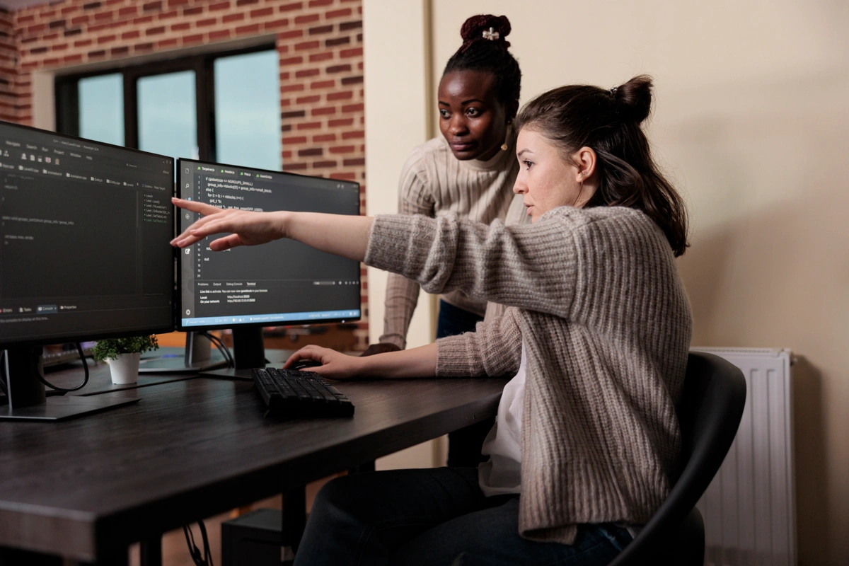 Caucasian network developer pointing at a computer screen while discussing high usage processing power with an African American programmer in a modern office setting.