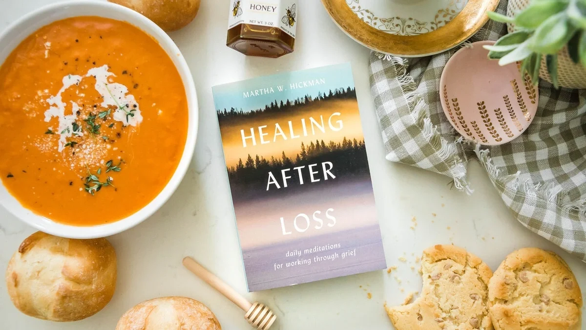 Close-up of a comforting arrangement featuring a bowl of tomato soup, a book titled Healing After Loss, rolls, cookies, a mini jar of honey with a wooden dipper, and a package of chamomile tea on a light surface.