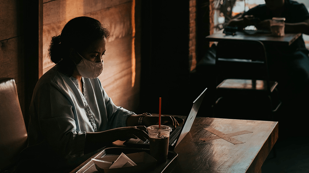 Woman with mask working on laptop in cafe