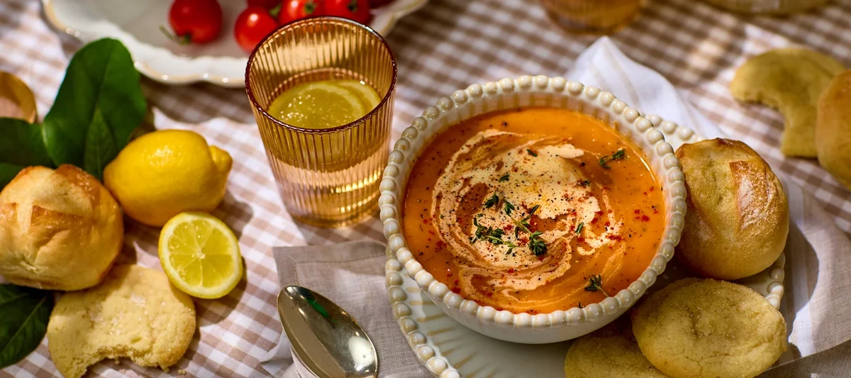A styled tabletop food scene featuring a bowl of creamy tomato-based soup garnished with herbs and a swirl of cream, surrounded by bread rolls, lemon halves, and a beverage on a checkered tablecloth.