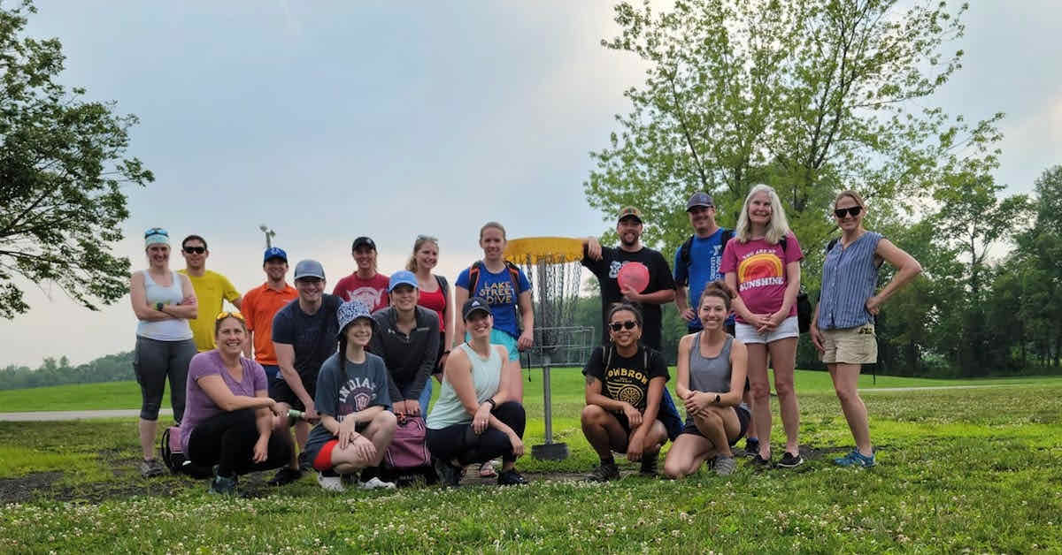 A group -- mostly women and some men-- pose for a photo around a disc golf basket