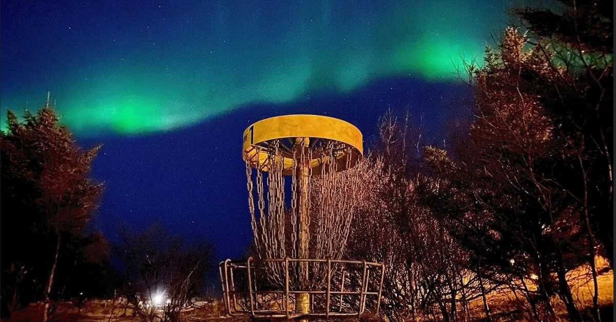 A disc golf basket in the dark with Northern Lights above it