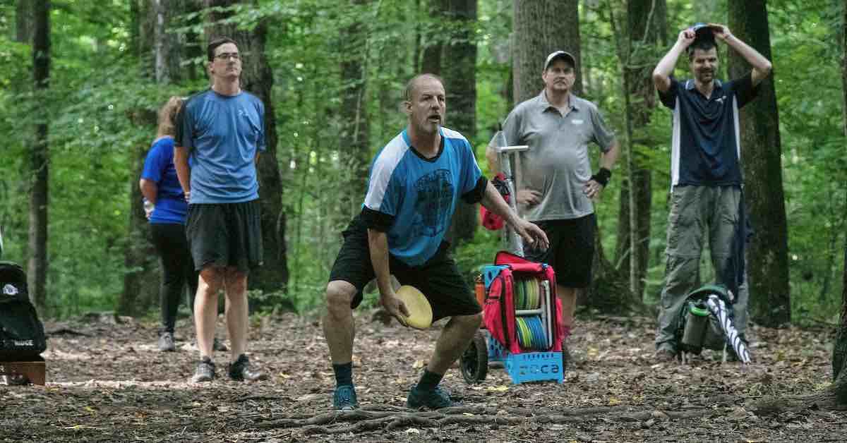 A disc golfer about to putt on a very wooded course while a small group stands respectfully behind
