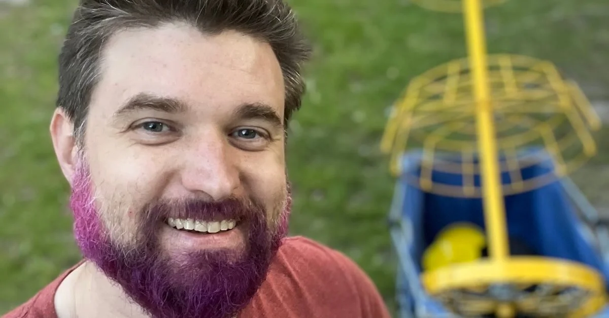 A man with a beard that has a bit of purple dye in it smiling in a close-up photo with disc golf basket in hand-pulled wagon behind