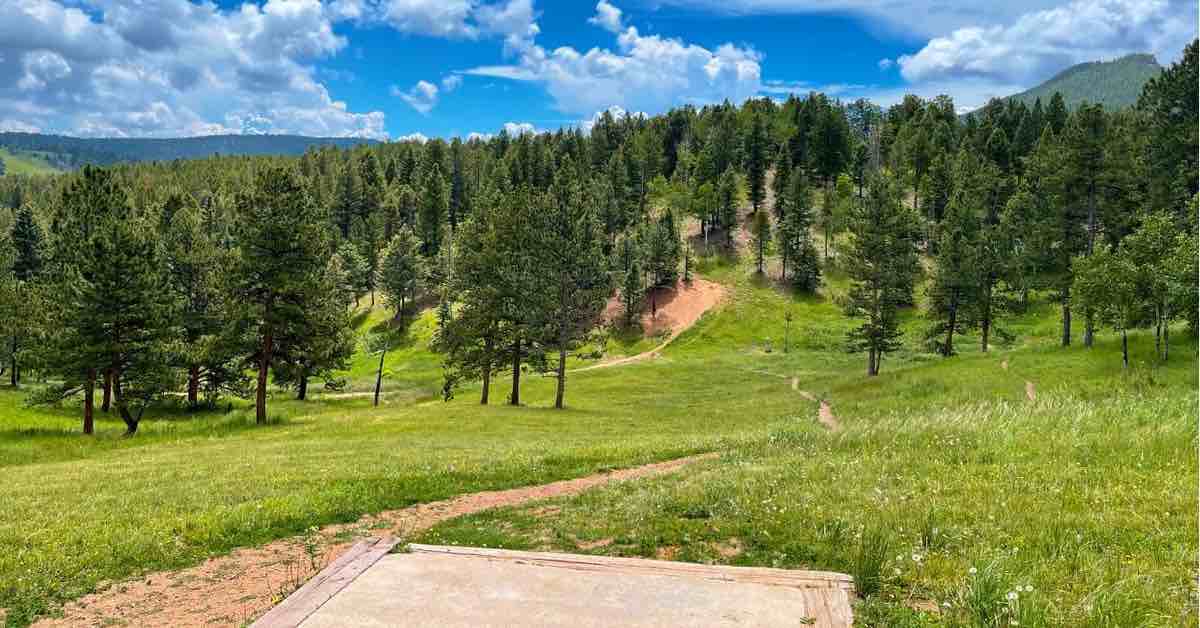 A concrete disc golf tee pad in an open grassy area with a view of evergreens, mountains, and blue sky