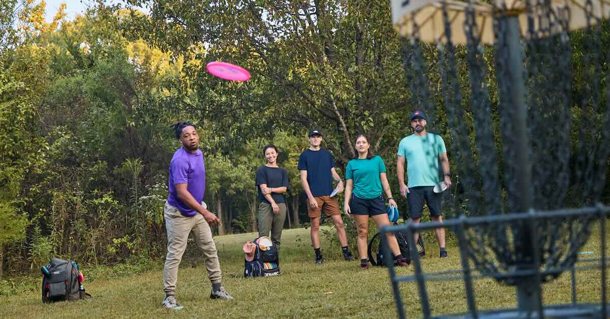 A man watches a disc golf glide toward a basket wwith four other players in the background looking on
