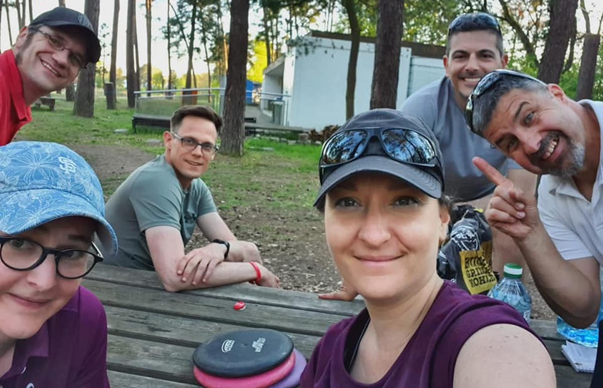 A group photo of two smiling women and four men at a picnic table outdoors with discs on table