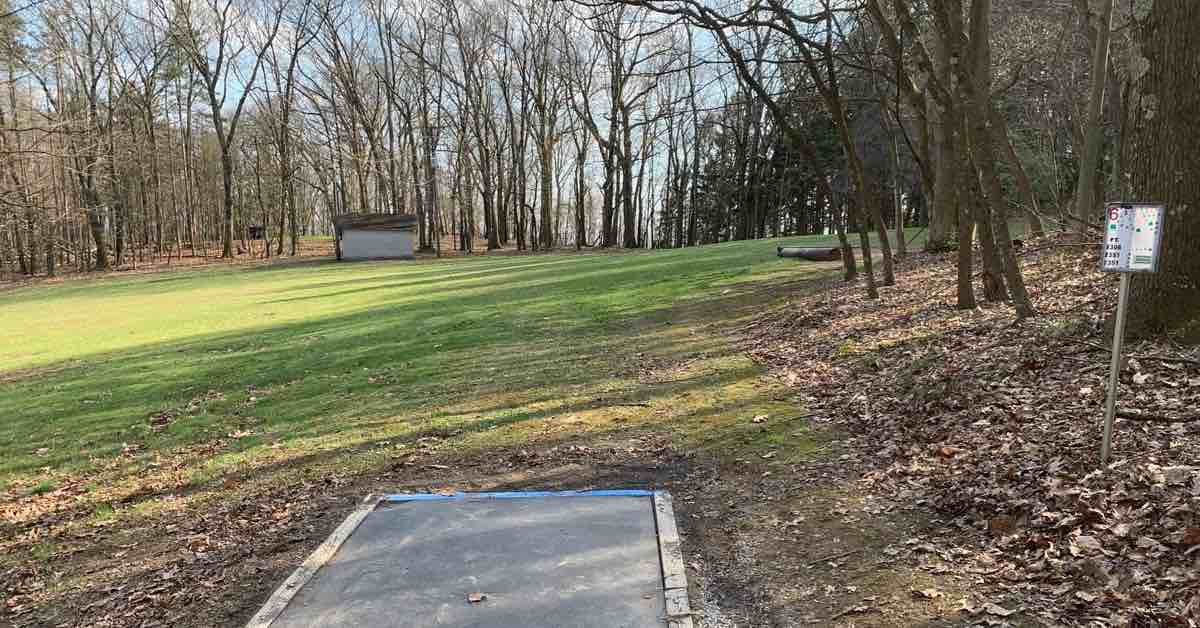 Rubber disc golf tee surface in open area leading to wooded fairway in hilly landscape